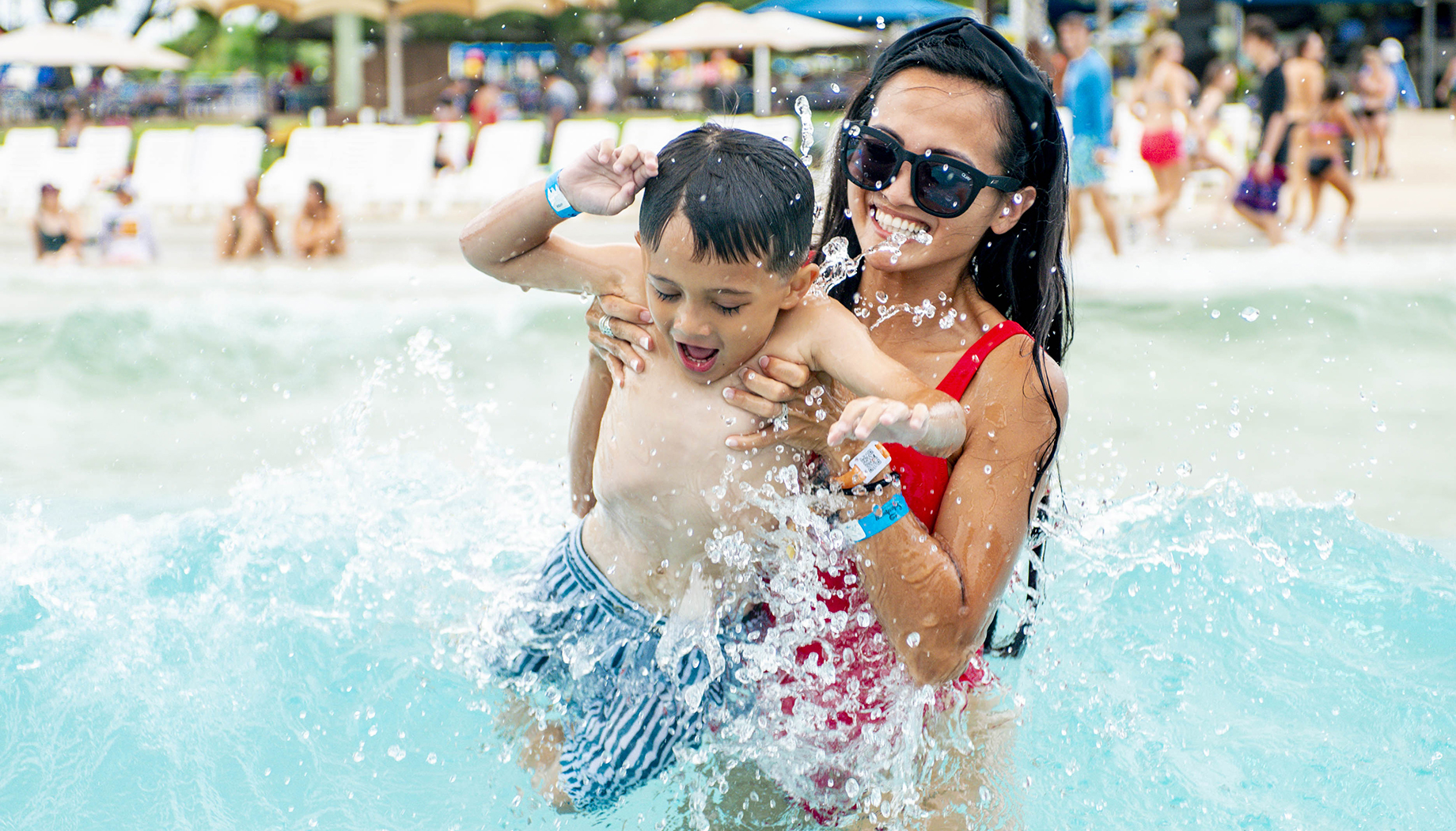 BLOG A woman in a red swimsuit lifts a young boy out of the water at a beach. Both are smiling, and there are other people and beach chairs in the background.