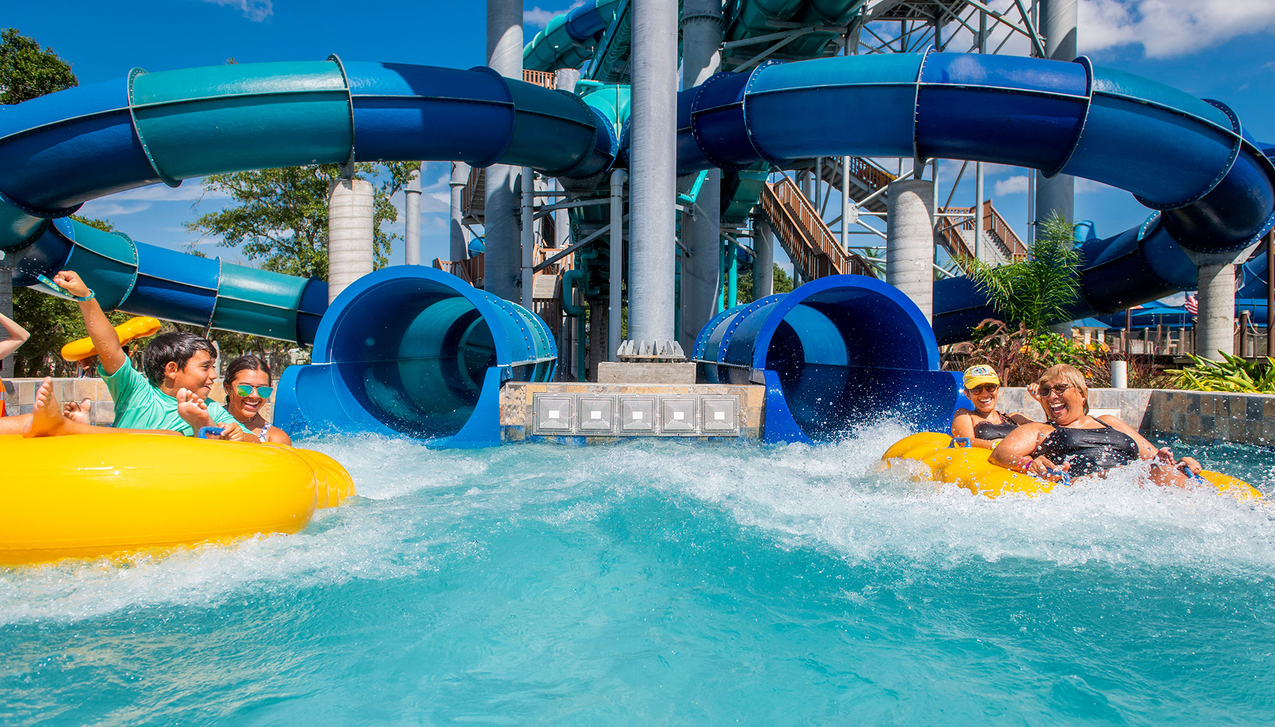 BLOG People riding yellow and blue inner tubes down a water slide at a water park with a backdrop of twisting water slide structures and clear blue sky.