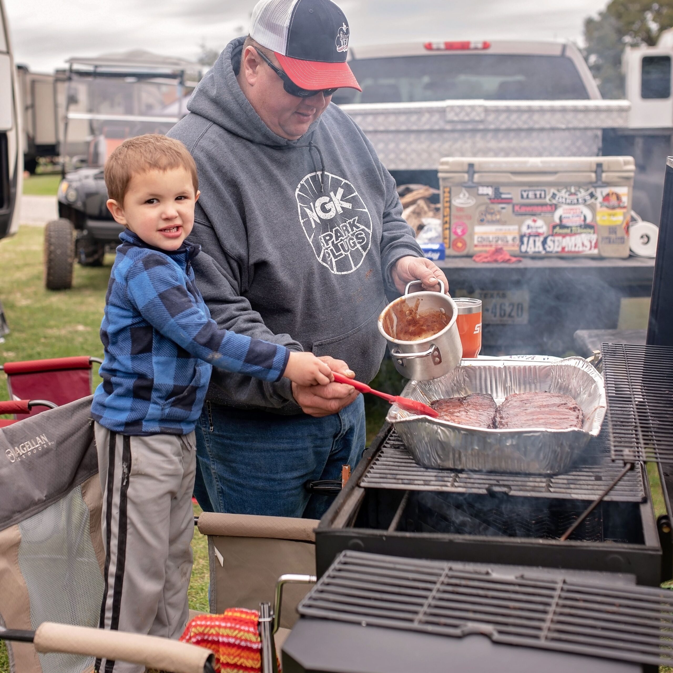 jAJttC5s (2) A man helps a young boy pour sauce over meat on a grilling barbecue during an outdoor event. The grill is set up next to a truck with various stickers.