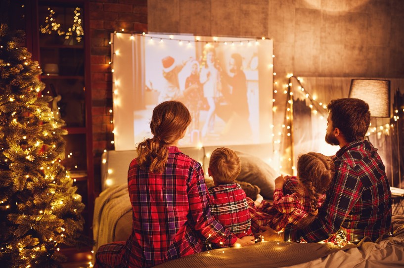 Screenshot 2023-12-11 144403 A family of four, dressed in matching plaid pajamas, watches a holiday movie projected on a screen in a cozy, warmly lit living room decorated with Christmas lights and a Christmas tree.